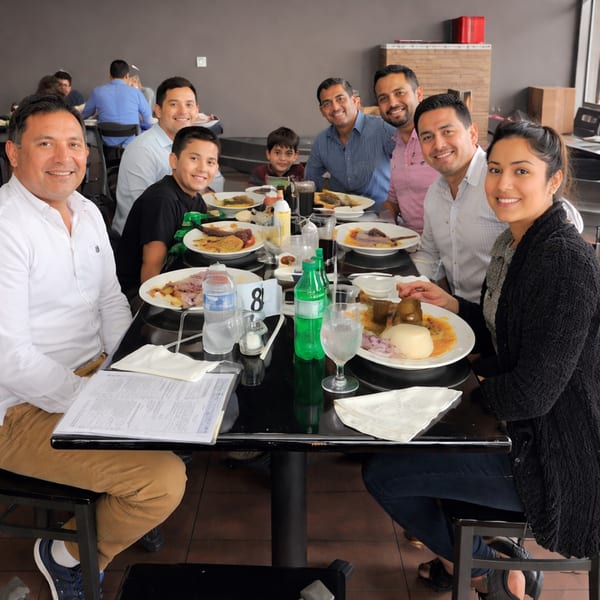 A group of smiling people, including men, women, and children, enjoying a meal at a restaurant table.