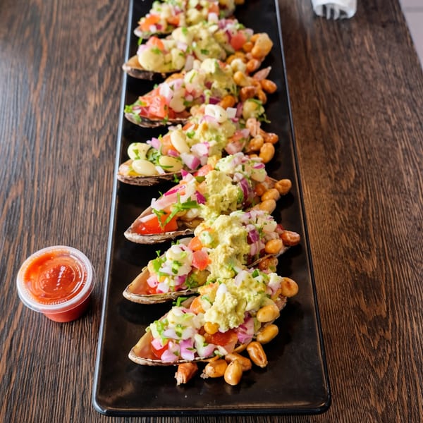 A black rectangular plate holds several mussels topped with salsa, guacamole, and corn nuts.
