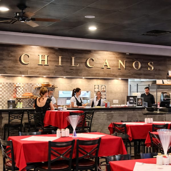 Restaurant interior with red tablecloths, black chairs, and staff behind a counter with "CHILCANOS" sign.