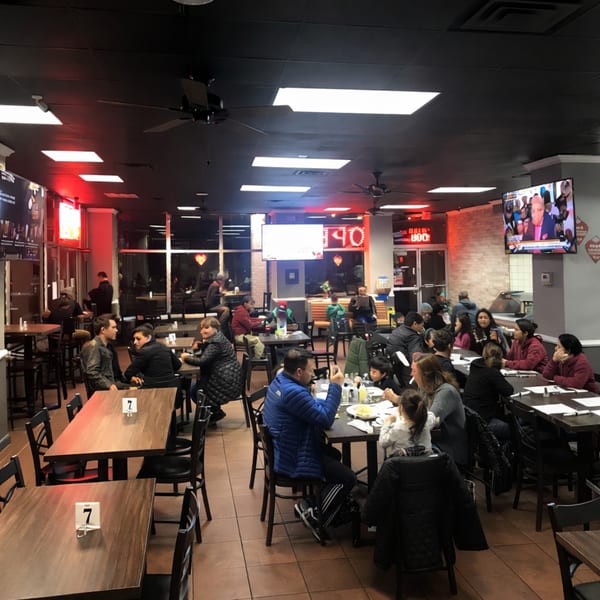 A busy restaurant interior with many patrons seated at tables, red neon lights, and TVs.