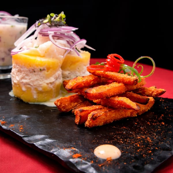 Peruvian causa with red onion and sweet potato fries on a black slate, with a drink in the background.