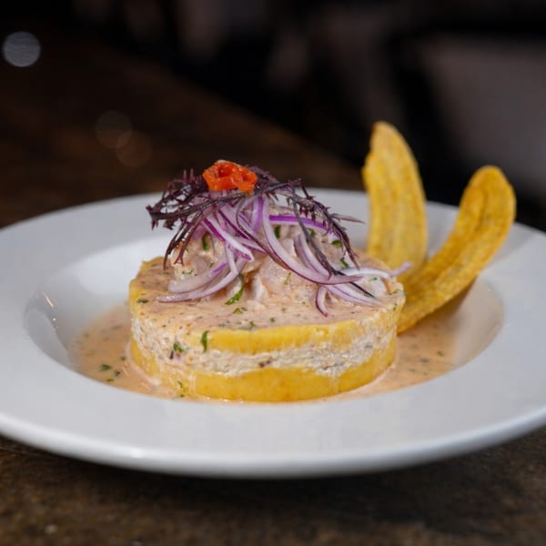 Peruvian causa with layered yellow potato, creamy filling, red onion, and plantain chips on a white plate.